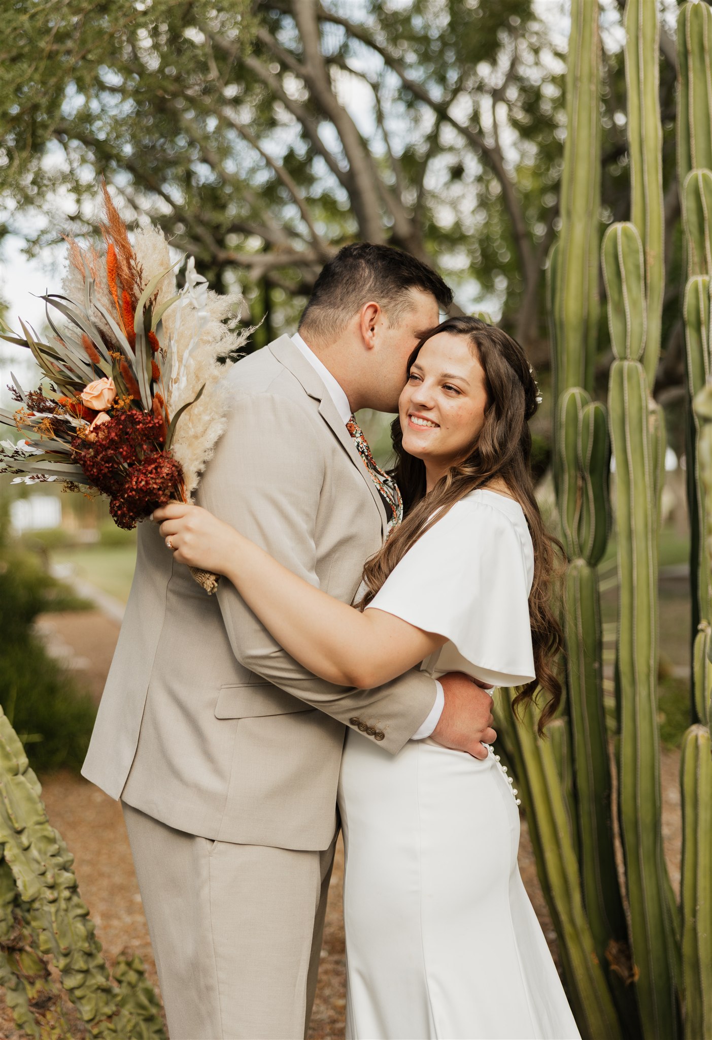 A bride smiles while holding her large boho bouquet and being kiss and hugged during their desert botanical gardens wedding