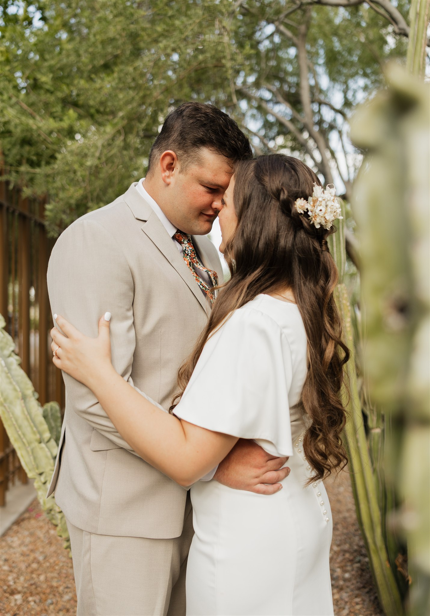 A groom in a tan suit snuggles his bride while touching foreheads during their desert botanical gardens wedding