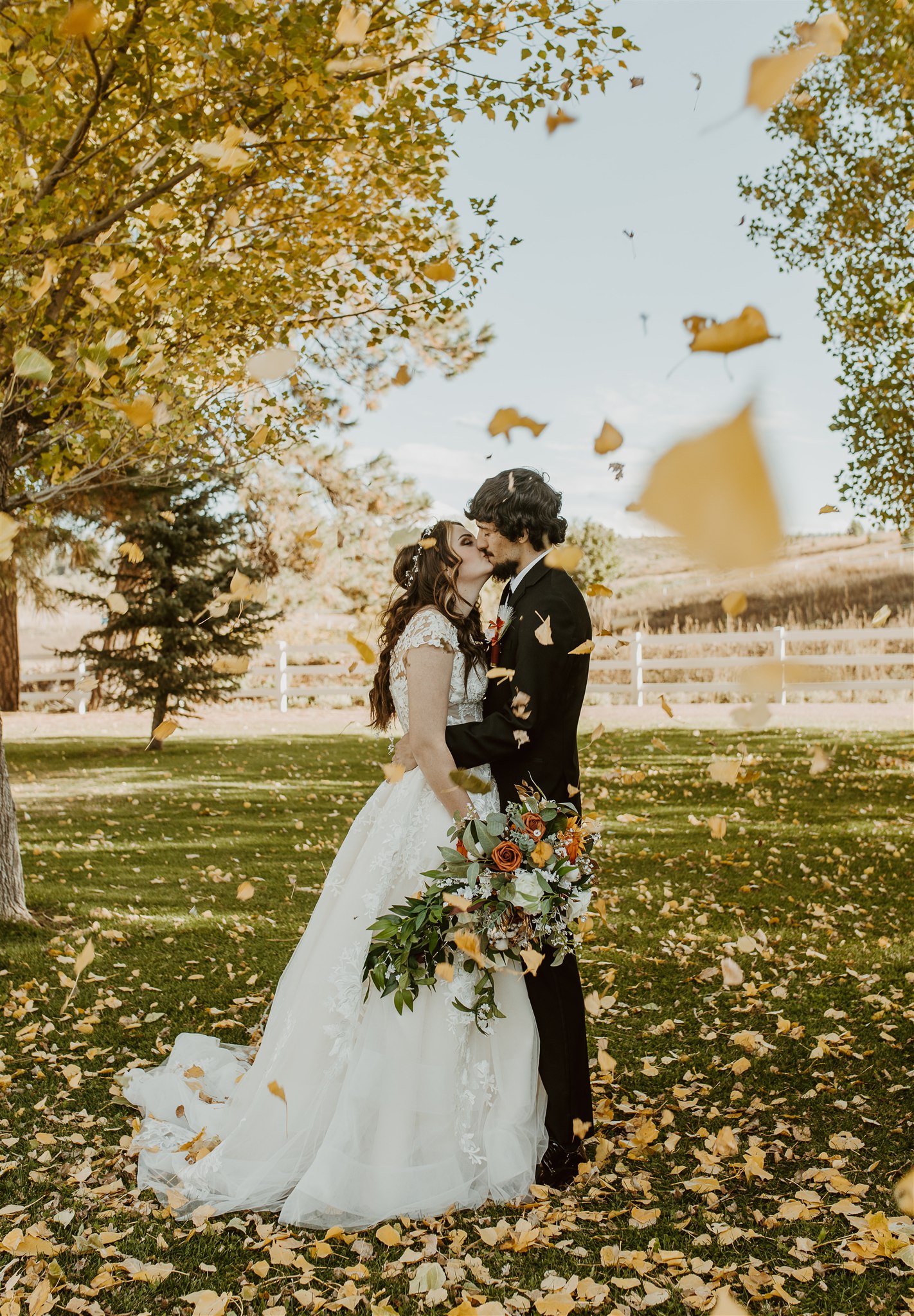 A bride and groom kiss as leaves fall around them in a lawn at sunset