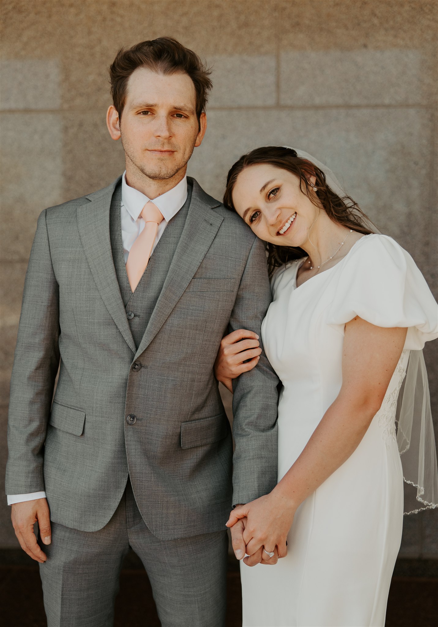 A bride rests on the shoulder of her groom while smiling and holding his hand in his grey suit with pink tie