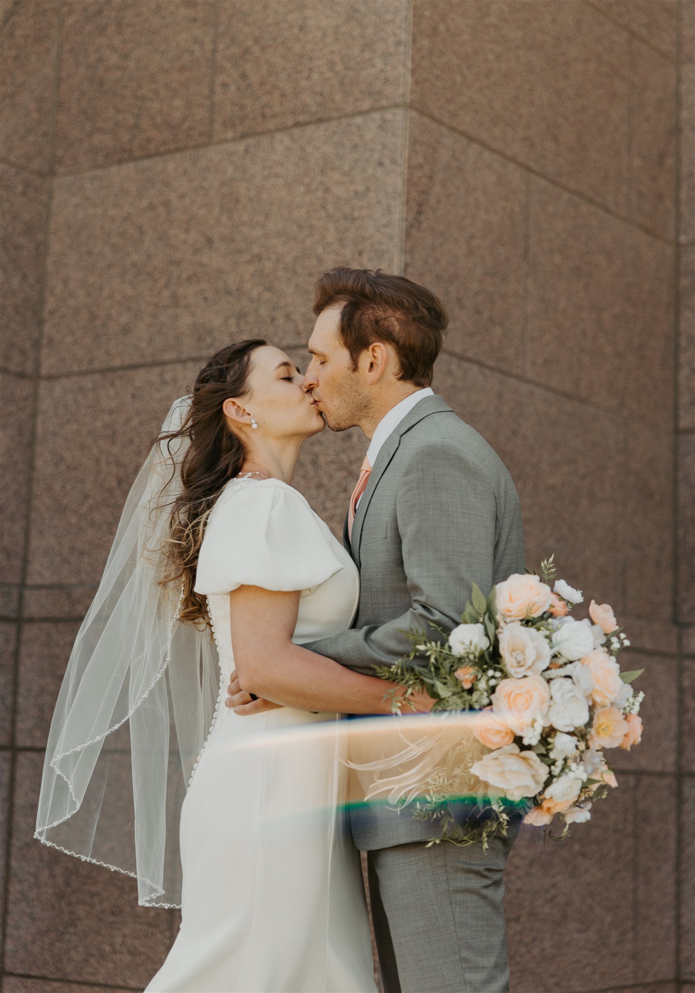 A bride and groom in a grey suit kiss next to a granite building with a sun flair during their day at The Icehouse wedding venue