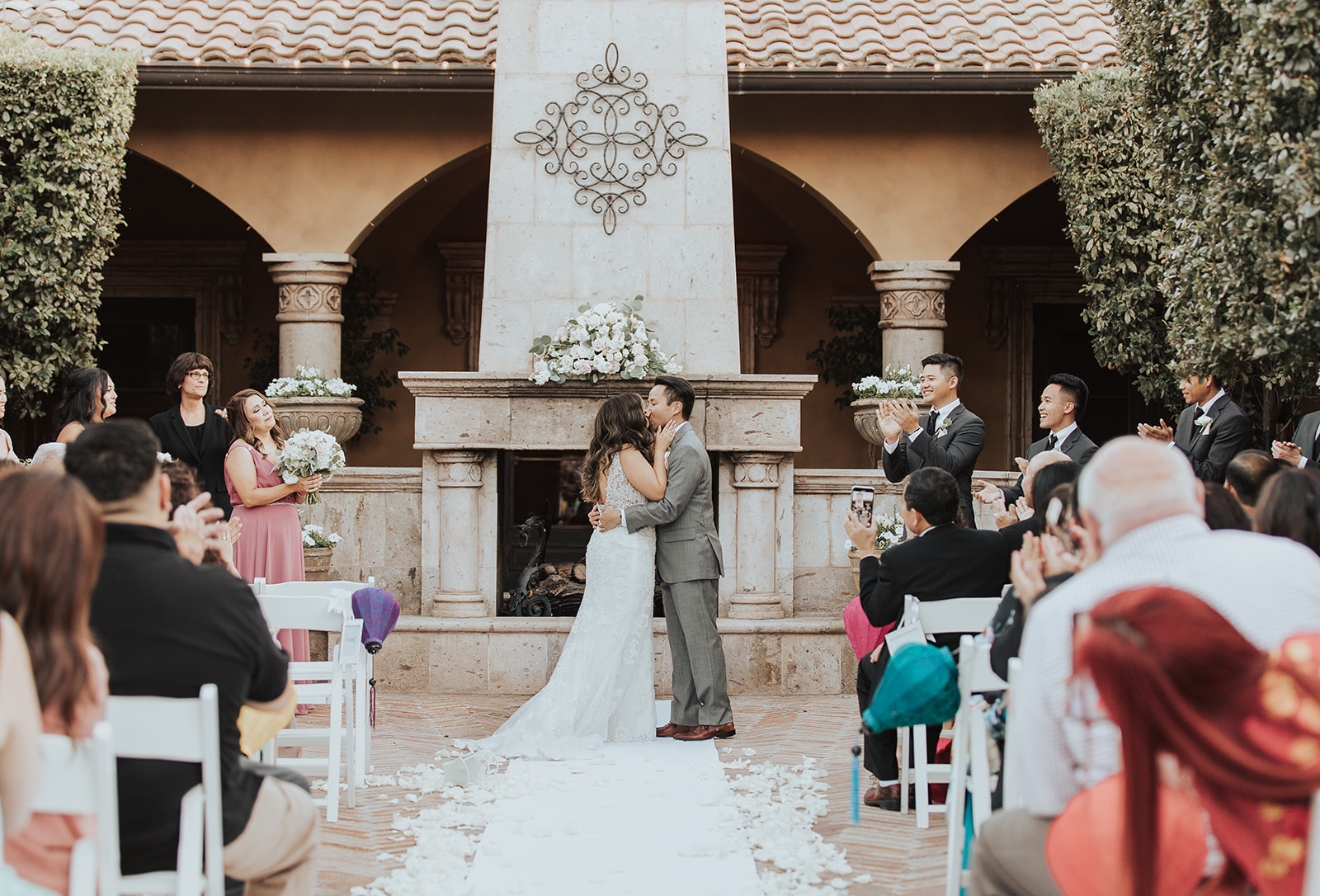 Newlyweds kiss in the gardens under a large white bouquet as guests clap to end their ceremony