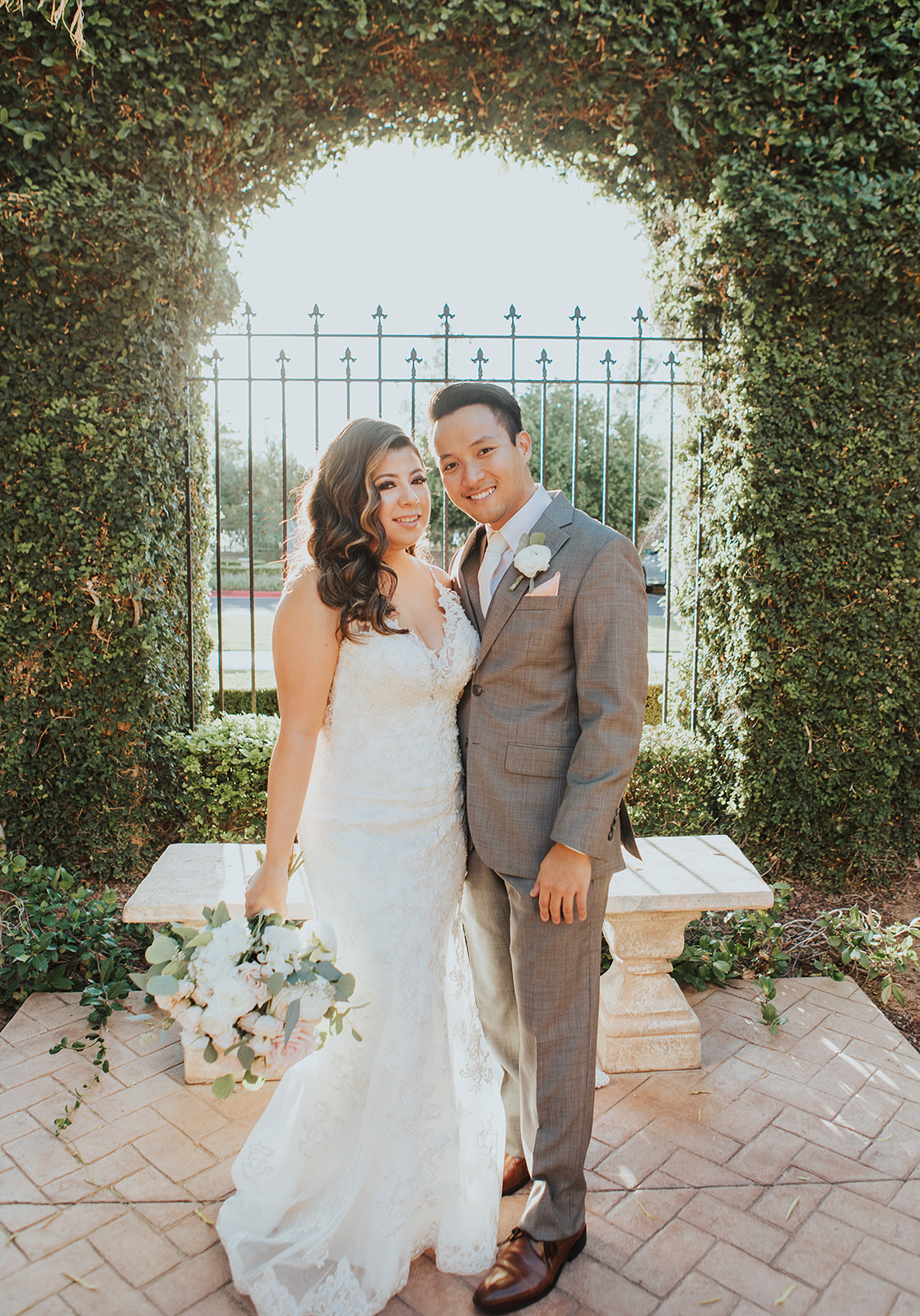 A bride smiles with her groom in grey under an ivy covered arch during their Villa Siena wedding
